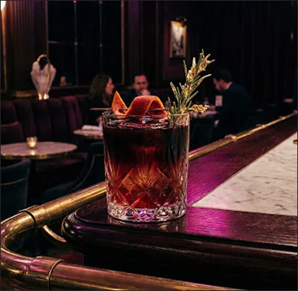 Un verre de cocktail ambré garni d'un zeste d'orange et d'un brin de romarin posé sur un comptoir de bar en bois poli