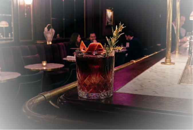 Cocktail artisanal ambré décoré d'un zeste d'orange et de romarin sur un comptoir de bar en marbre dans une ambiance tamisée