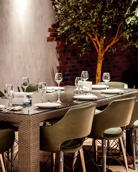 Une grande table de restaurant élégamment dressée avec des verres à vin, des assiettes et des chaises modernes près d'un mur en briques et d'un arbre décoratif